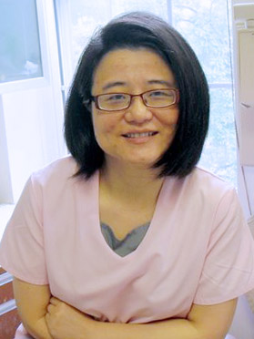 A woman wearing glasses, a short-sleeved shirt, and a lanyard around her neck, posing in front of a window with blinds partially drawn.
