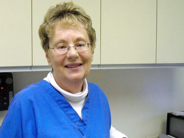 The image shows a woman in a blue uniform, smiling at the camera, standing in what appears to be a kitchen or break room area with white cabinets and a microwave.
