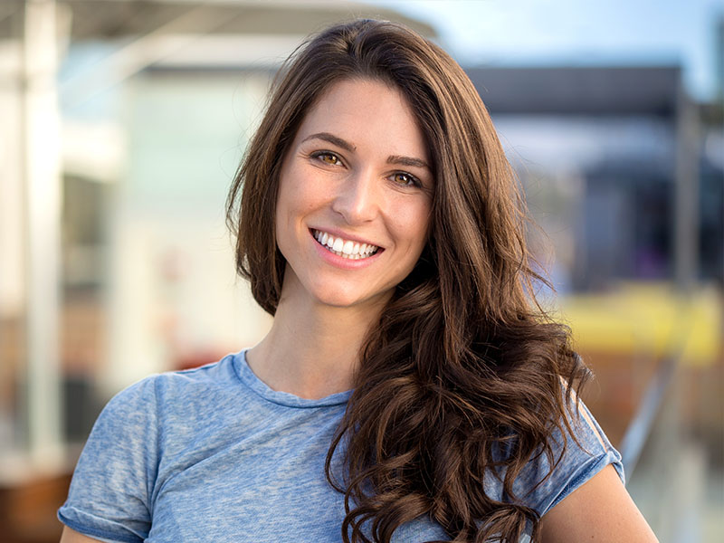 The image is a portrait of a woman with long brown hair, smiling at the camera. She has a light complexion and appears to be outdoors during daylight hours. The woman is wearing a casual top with a visible logo on it, suggesting she might be affiliated with a brand or organization. Her pose is relaxed, with her left hand resting on her hip and her right arm slightly extended from her body. She is standing in front of what seems to be a building with large windows, which could indicate a commercial or public space. The sky is visible in the background, suggesting an open-air setting.