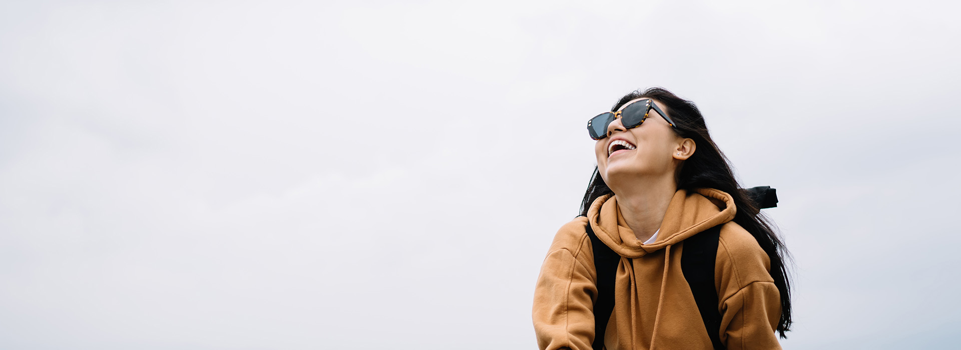 The image features a woman standing outdoors, smiling and looking upwards towards the camera. She is wearing sunglasses, a backpack, and a hoodie with a light-colored background.