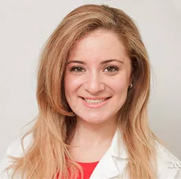 Female dental professional in white coat smiling at camera against neutral background.